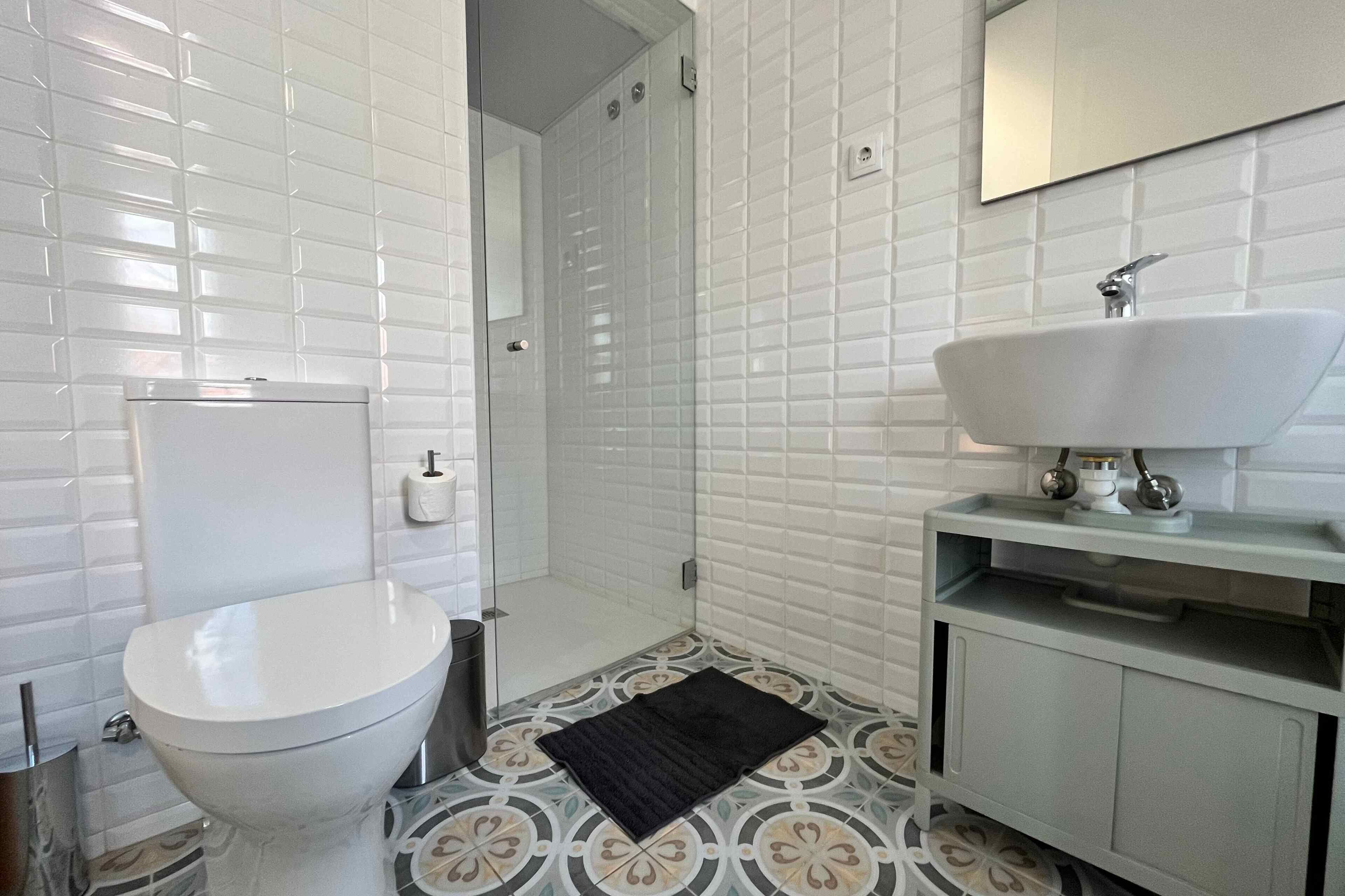 A bathroom with white tiled walls, a white toilet, a sink with a mirror above it, and patterned floor tiles.
