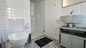 Un baño con paredes de azulejos blancos, un inodoro blanco, un lavamanos con espejo y baldosas de suelo estampadas.