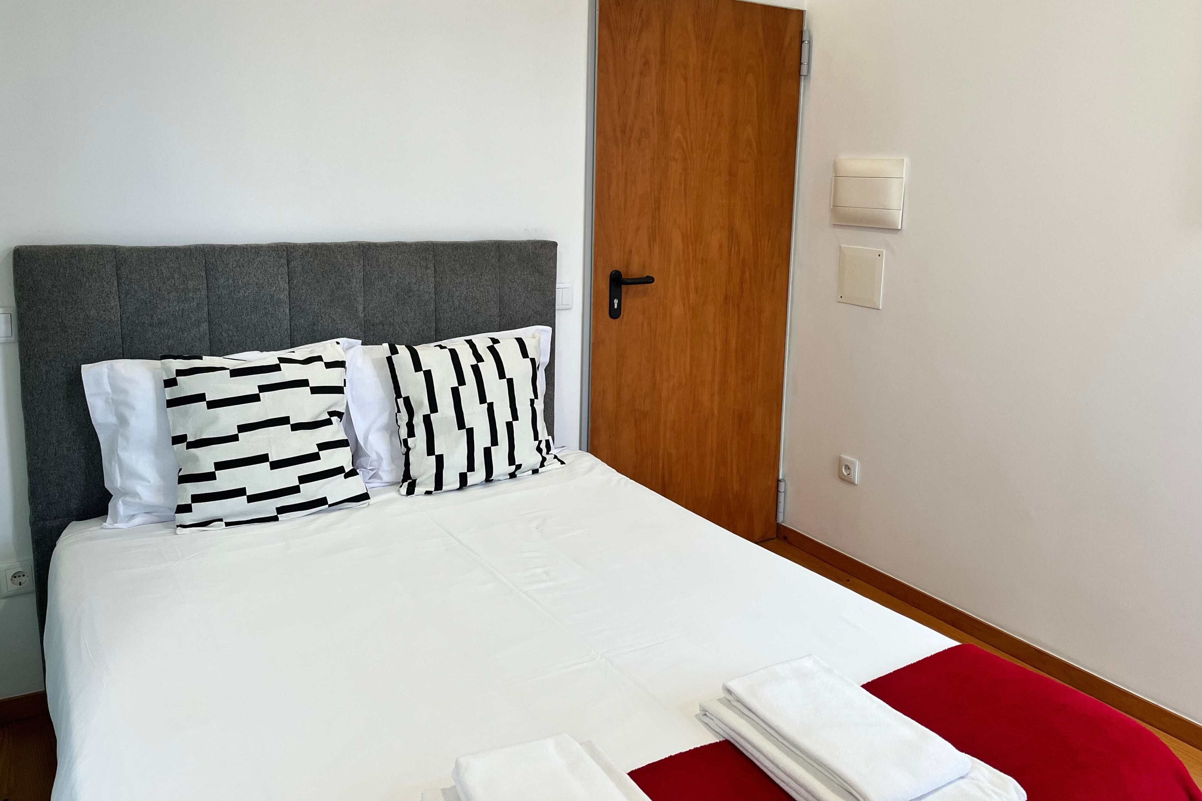 A neatly made bed with white bedding and two patterned pillows, a wooden door, and a red accent on the floor.