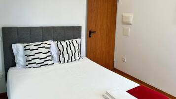 A neatly made bed with white bedding and two patterned pillows, a wooden door, and a red accent on the floor.