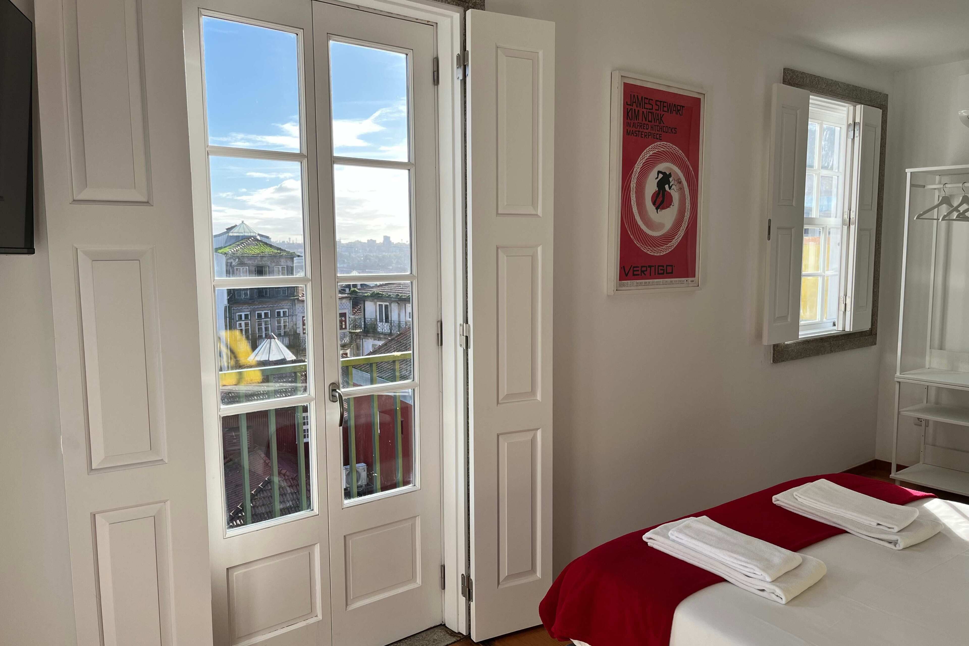 Une chambre d’hôtel avec une grande fenêtre, un lit avec du linge de lit blanc, un plaid rouge et une affiche accrochée au mur.