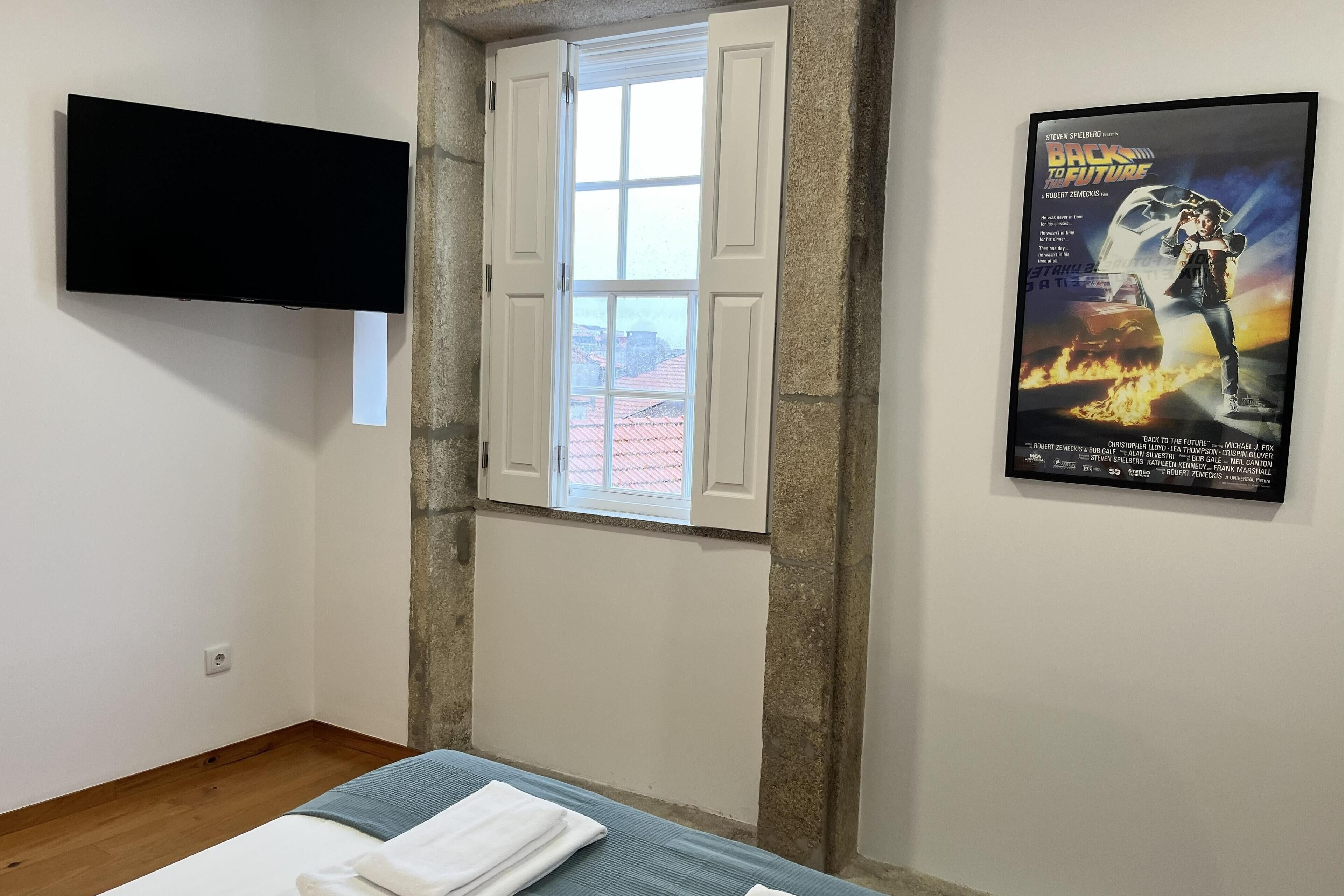 A hotel room with a bed, a TV mounted on the wall, a window with open shutters, and a movie poster on the wall.