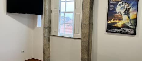 A hotel room with a bed, a TV mounted on the wall, a window with open shutters, and a movie poster on the wall.