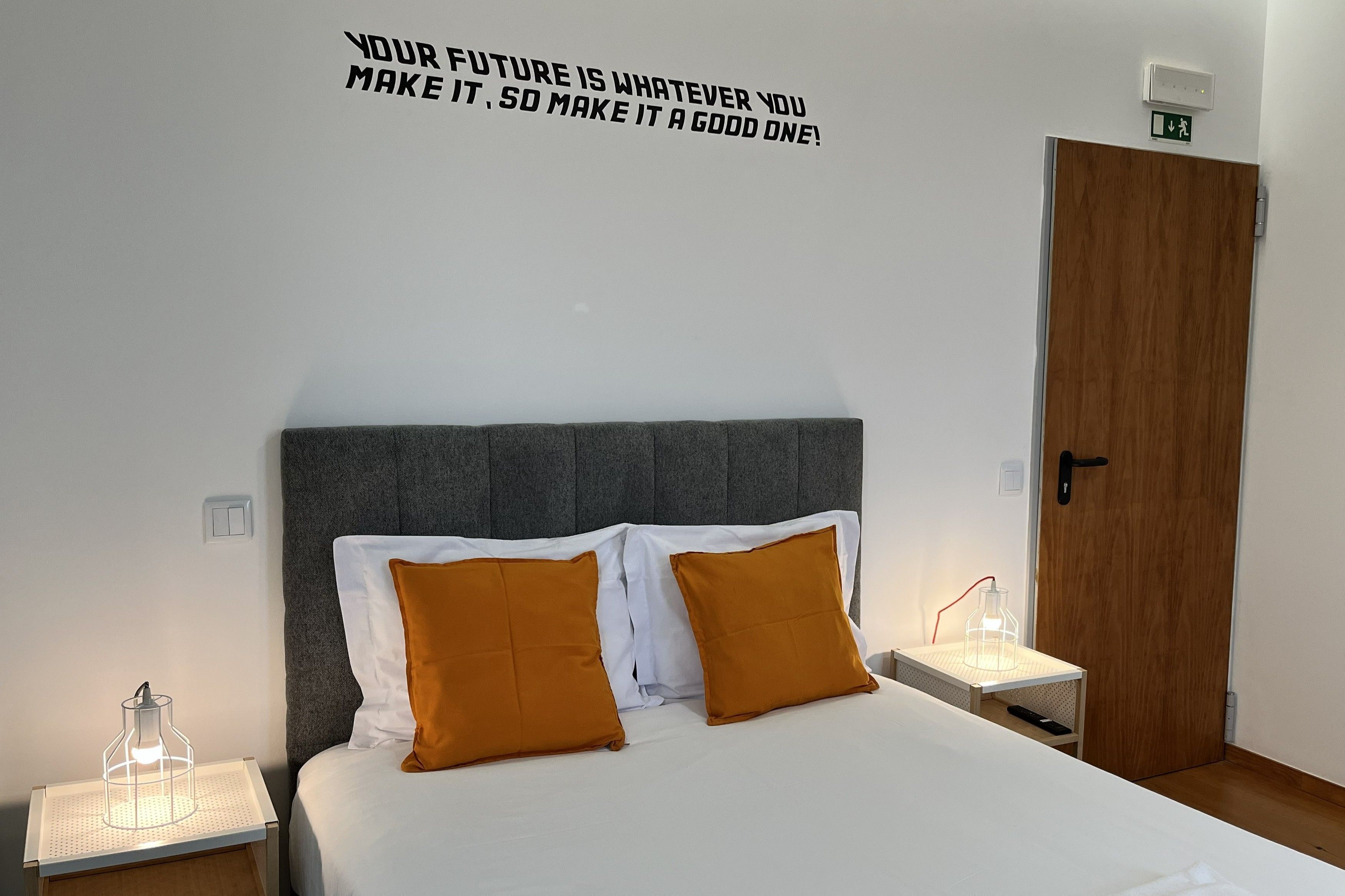 Una cama bien hecha con almohadas blancas y mostaza, un cabecero tapizado en gris y una cita motivacional en la pared.
