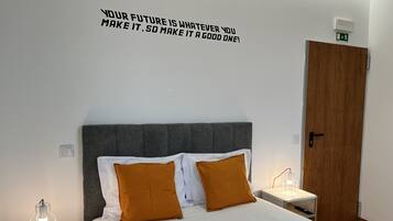 Una cama bien hecha con almohadas blancas y mostaza, un cabecero tapizado en gris y una cita motivacional en la pared.