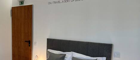 Quarto de hotel com cama, duas luminárias de cabeceira, porta de madeira e um letreiro na parede.