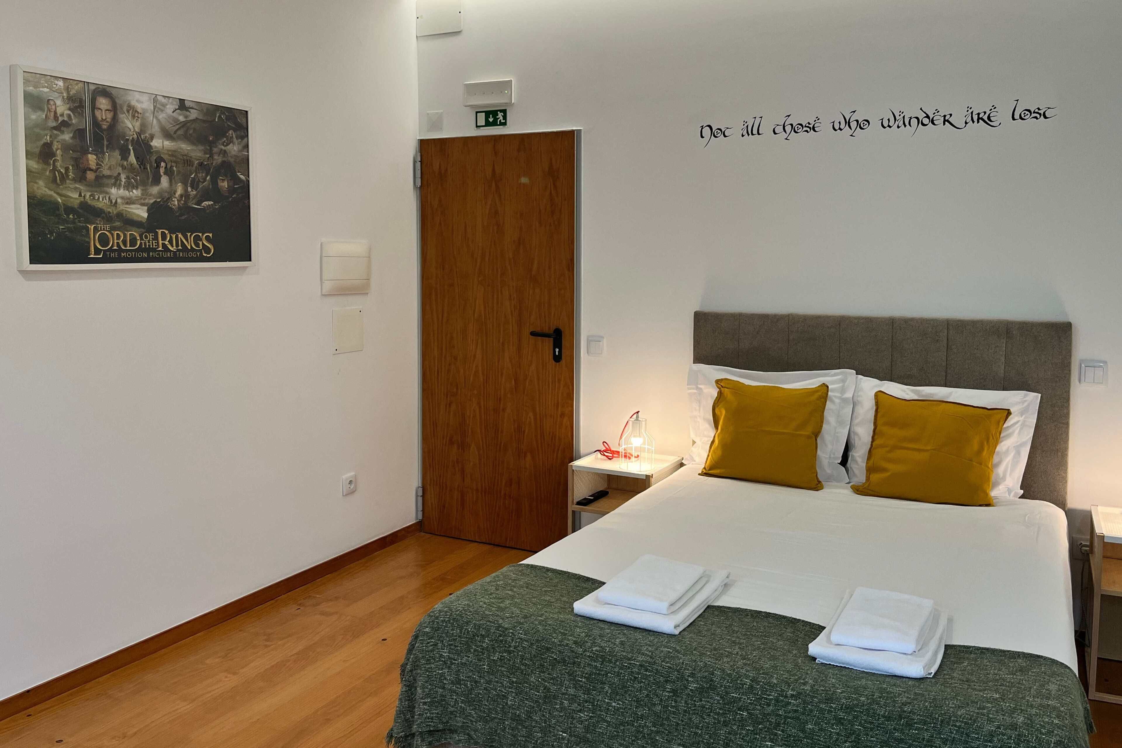 A hotel room with a bed, a poster of 'The Lord of the Rings' trilogy, a wall quote, and a wooden door.