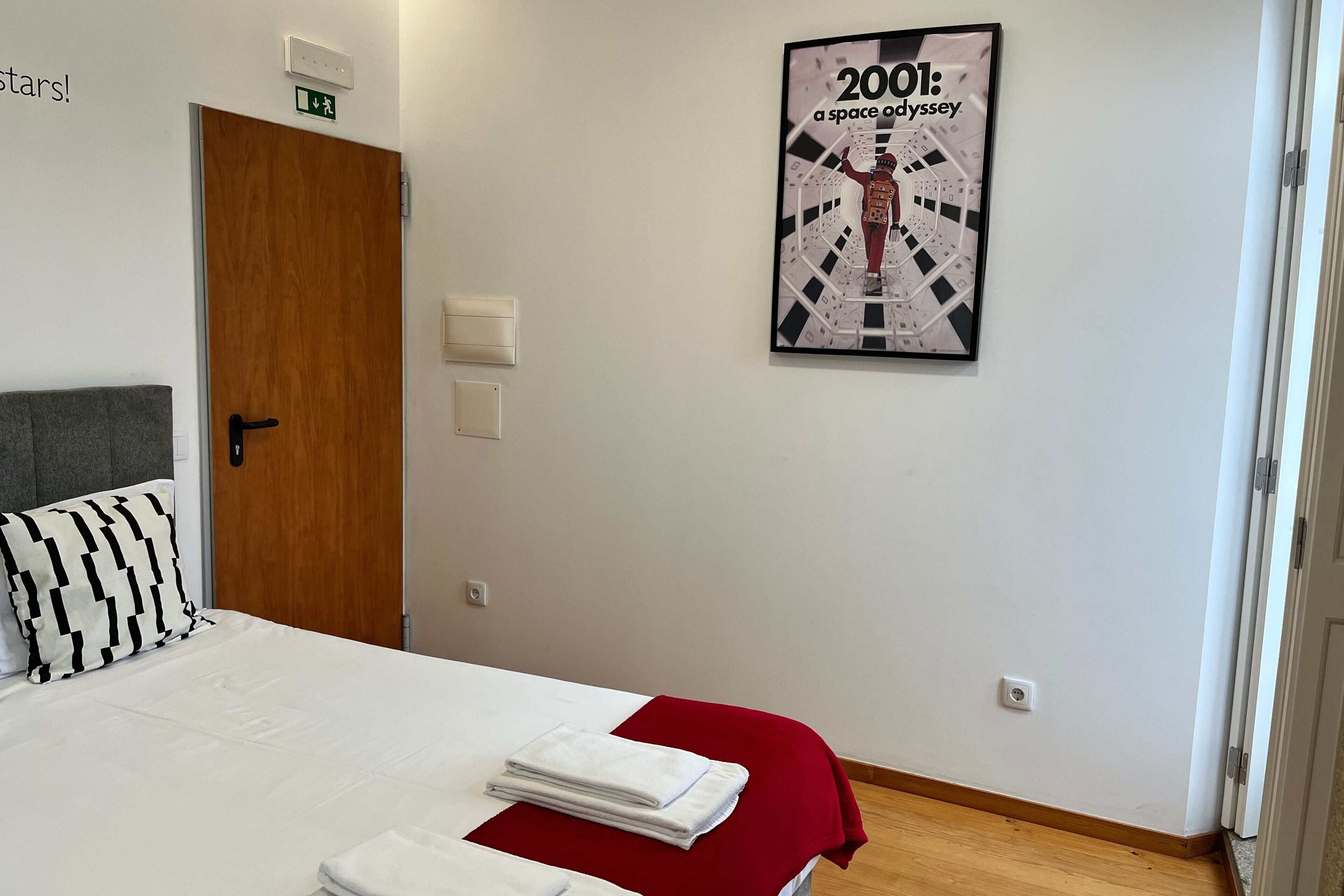 Quarto de hotel com cama, manta vermelha, pôster emoldurado, porta de madeira e cabeceira cinza.