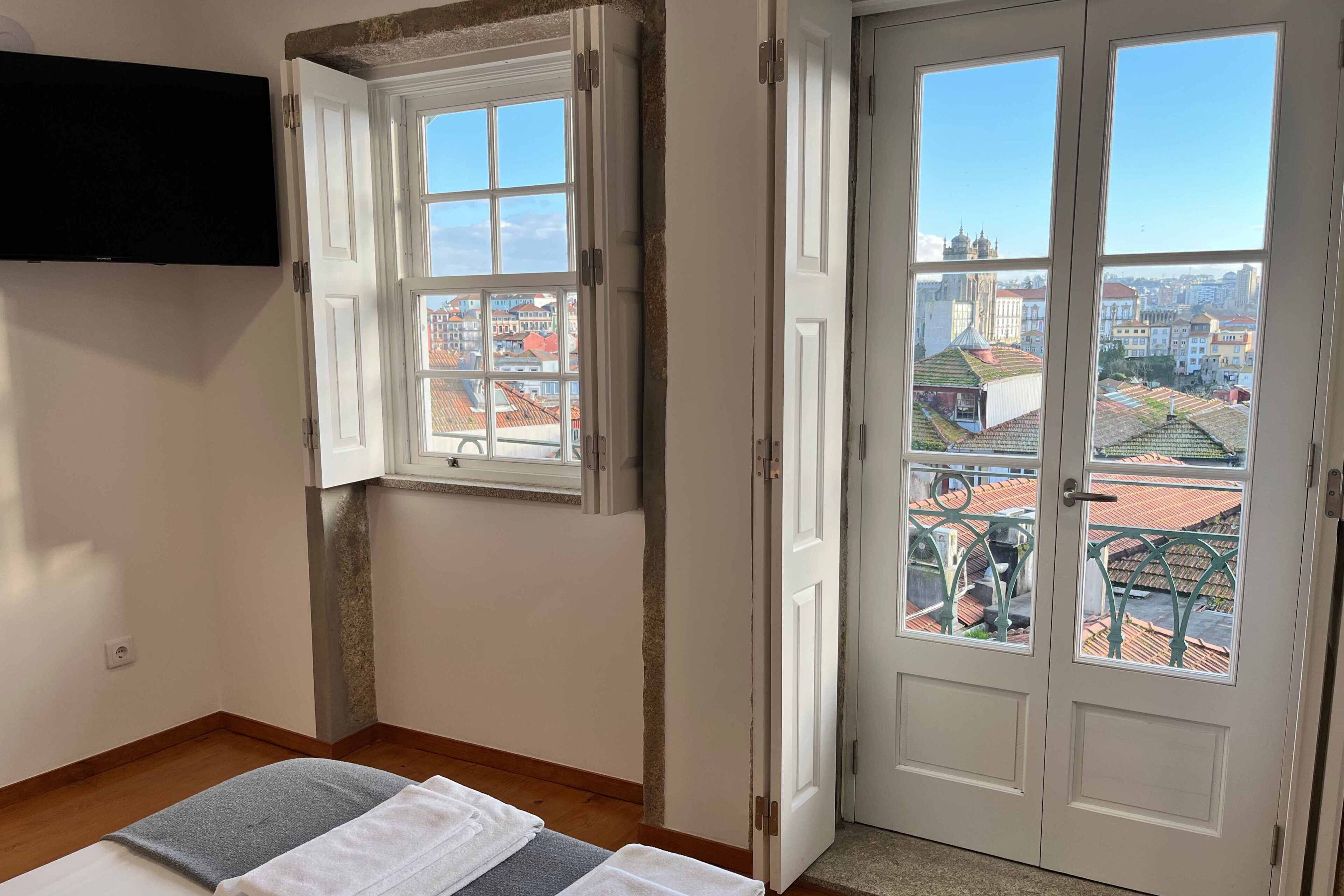 A room with a bed, a TV mounted on the wall, a window with a view of the city, and a door leading outside.