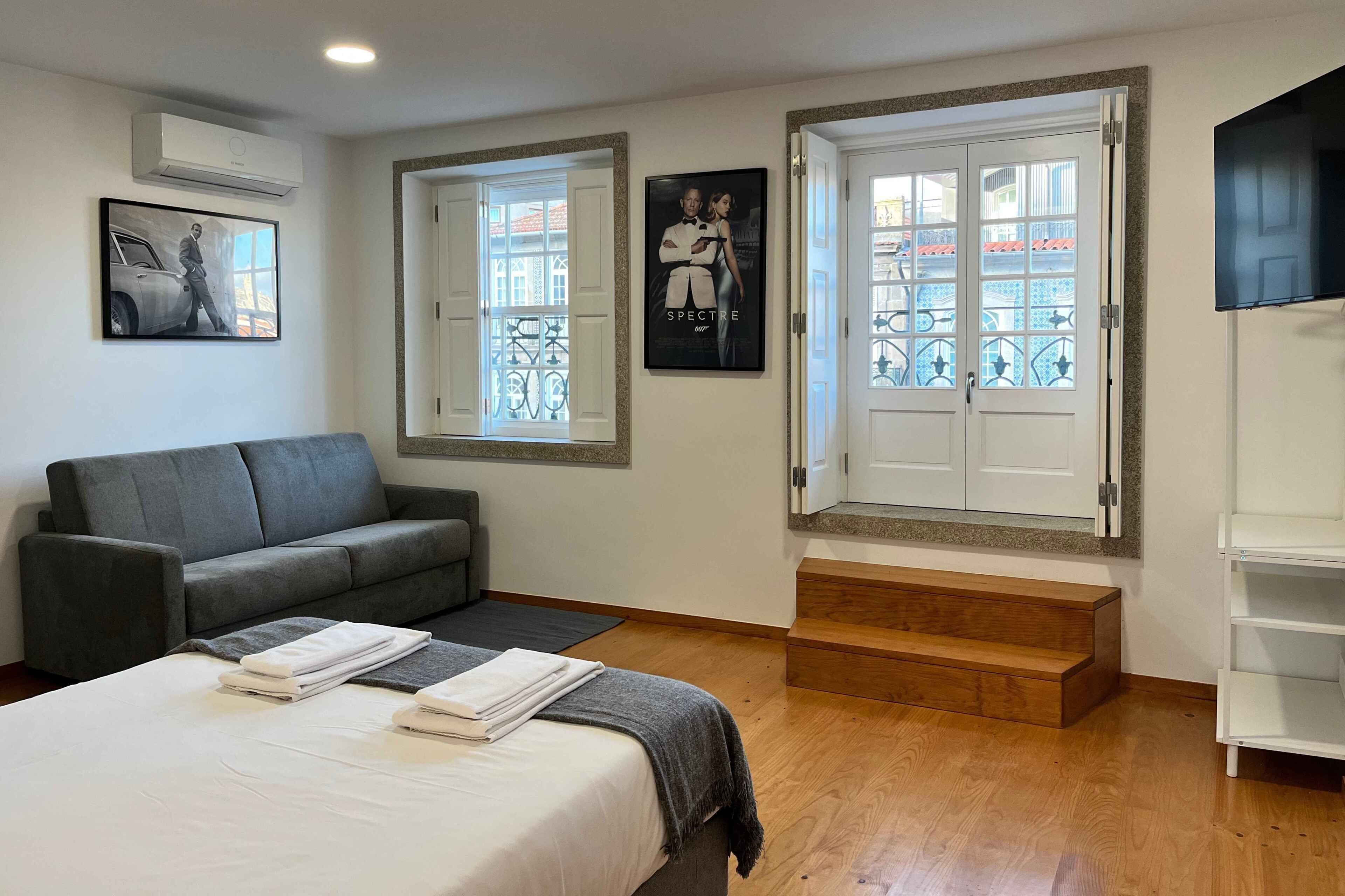 A hotel room with a bed, a grey sofa, a staircase, and two windows with a view of a balcony.