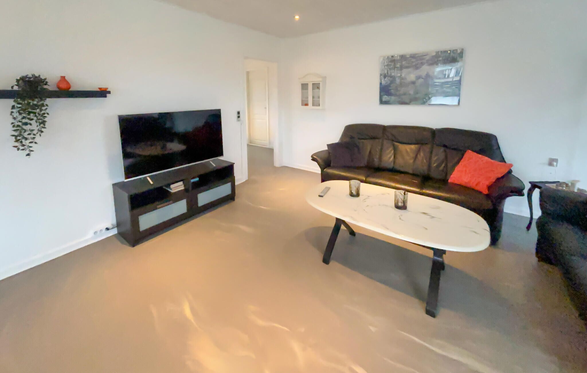 A living room with a flat-screen TV on a black stand, a white coffee table, a black leather sofa with a red pillow, and a small plant on a shelf.