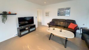 Ein Wohnzimmer mit einem Flachbildfernseher auf einem schwarzen Standfuß, einem weißen Couchtisch, einer schwarzen Ledersofa mit einem roten Kissen und einer kleinen Pflanze auf einem Regal.