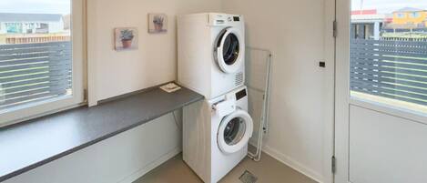 A laundry room with two stacked washing machines, a drying rack, and a window with blinds.