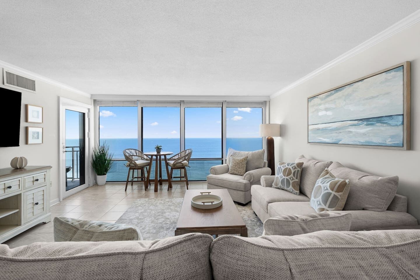 A spacious living room with a large window overlooking the sea, a comfortable sofa set, a dining area with chairs, and a flat-screen TV mounted on the wall.