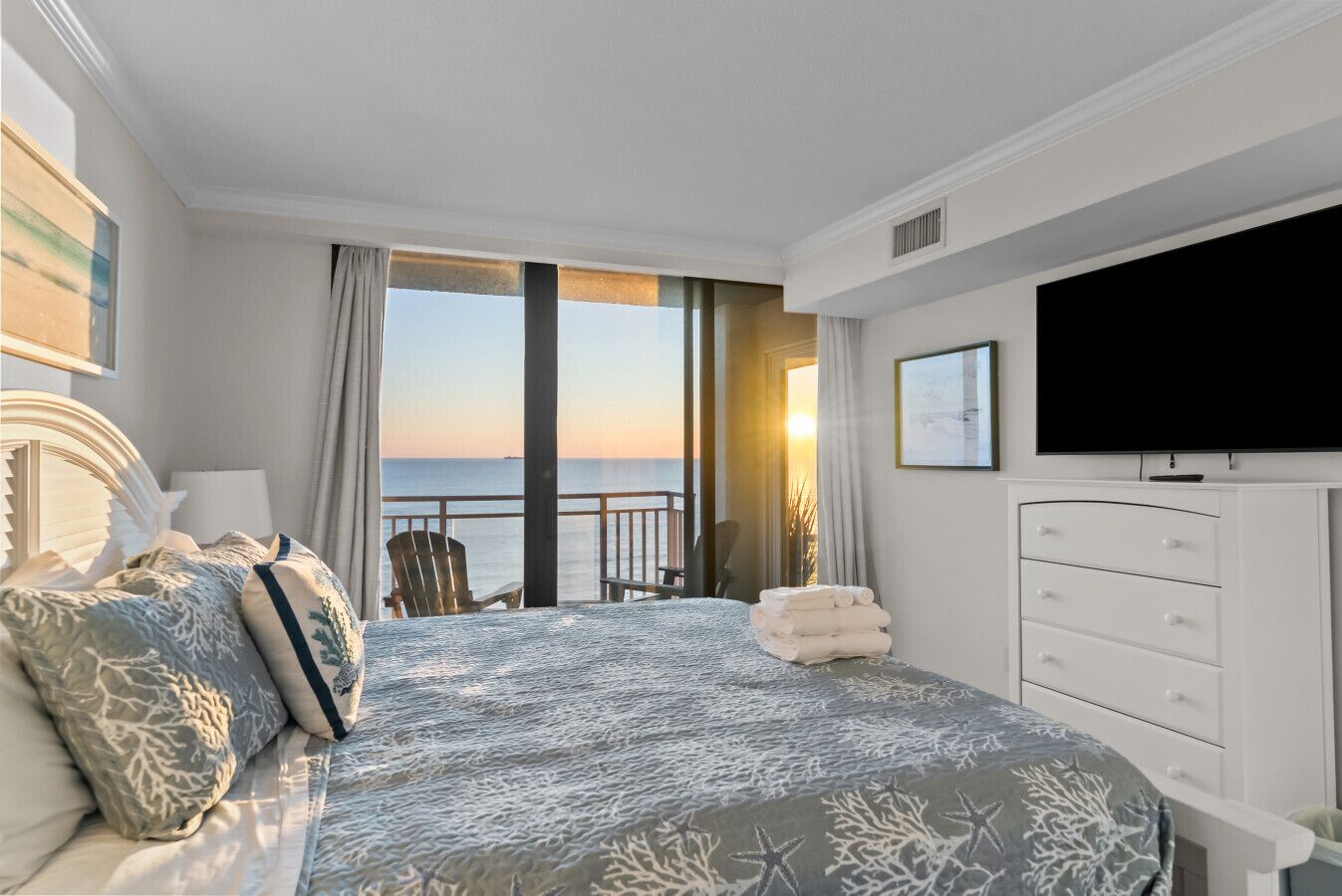 A hotel room with a large bed, a TV, a dresser, and a balcony with a view of the ocean.