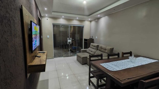 A modern living room with a large TV, a dining table with chairs, a sofa, and a glass door leading to a balcony. - 3-Bedroom House with Garage and BBQ Area (Casimiro de Abreu)