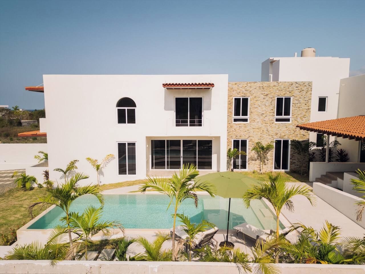 A modern two-story house with a swimming pool, surrounded by palm trees and a clear sky.