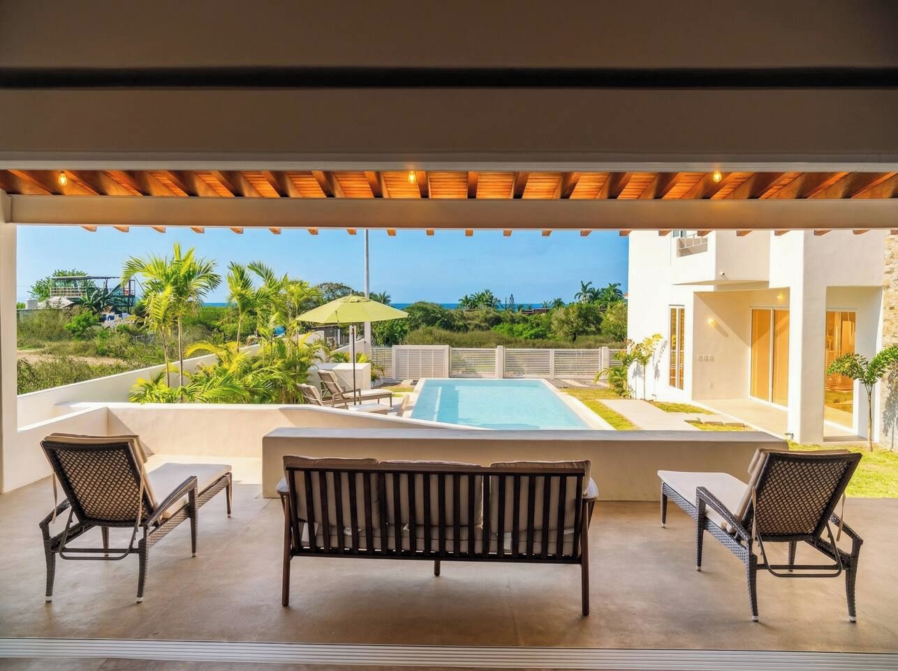 Un espace aménagé au bord de la piscine, avec un banc et des chaises, donnant sur une piscine, et avec un bâtiment visible au loin.