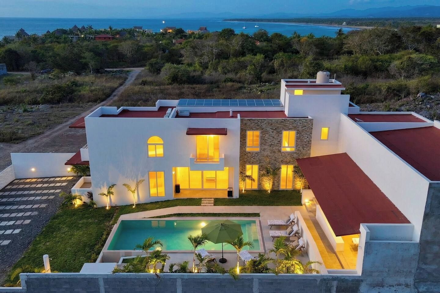 A modern two-story house with a swimming pool, surrounded by greenery and overlooking a coastal area.
