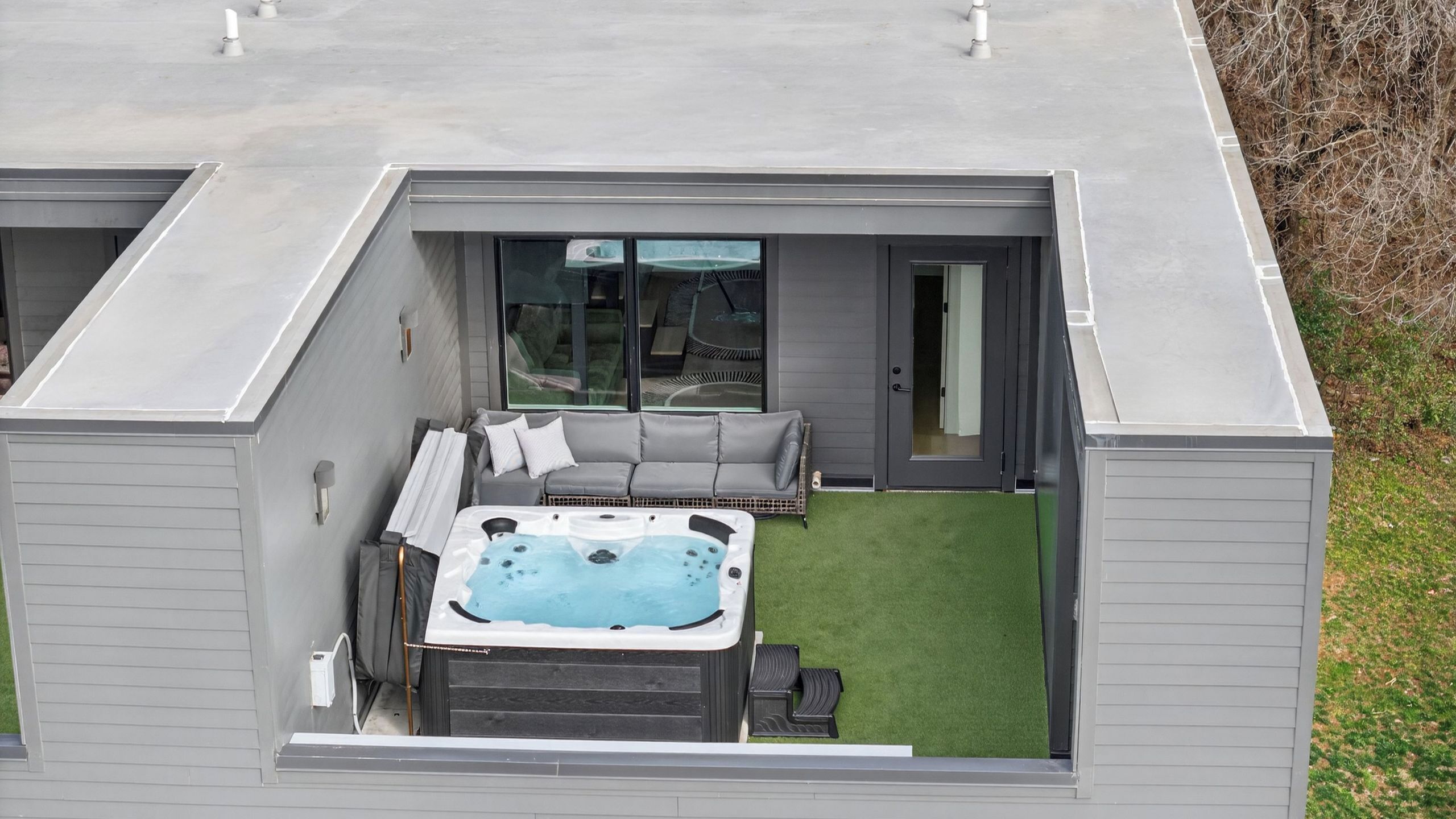 Eine moderne Außenpatio mit Whirlpool, einem grauen Sofa und einer Glastür zu einem Innenbereich.