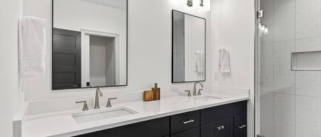 A modern bathroom with a white vanity, two sinks, a large mirror, and a shower area.
