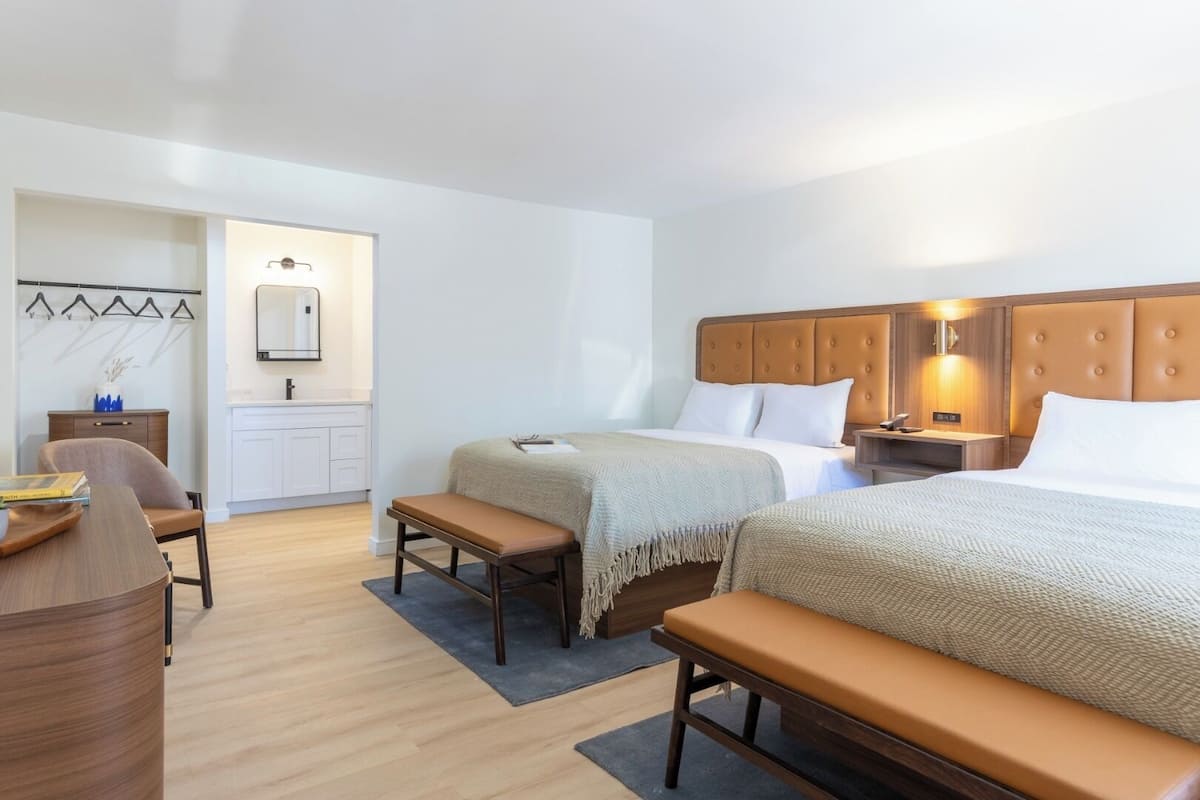 A hotel room with two beds, a desk, a chair, a bench, a small kitchenette, and a bathroom.