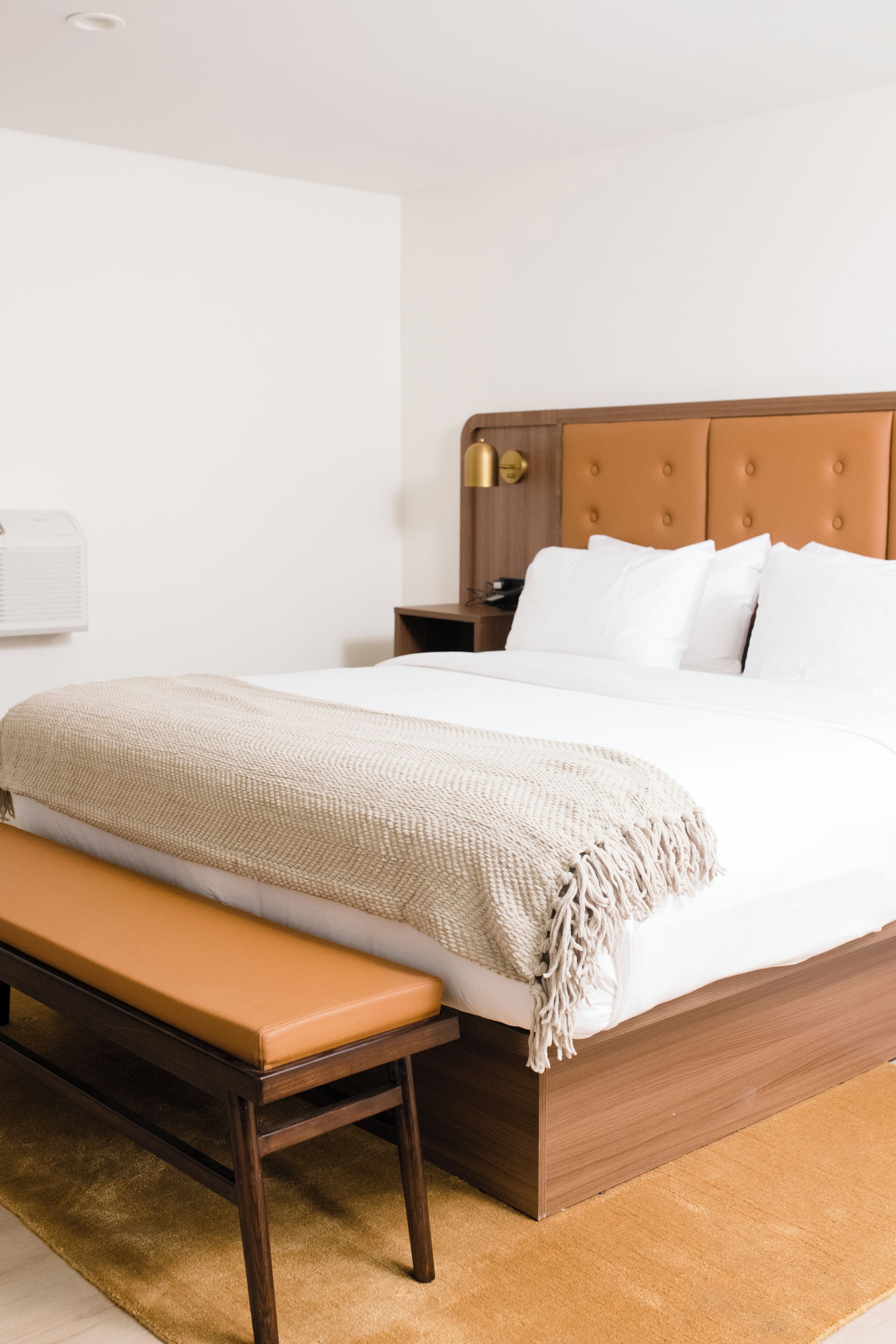 A neatly made bed with a wooden headboard, a bench at the foot of the bed, and a bedside table with a lamp.