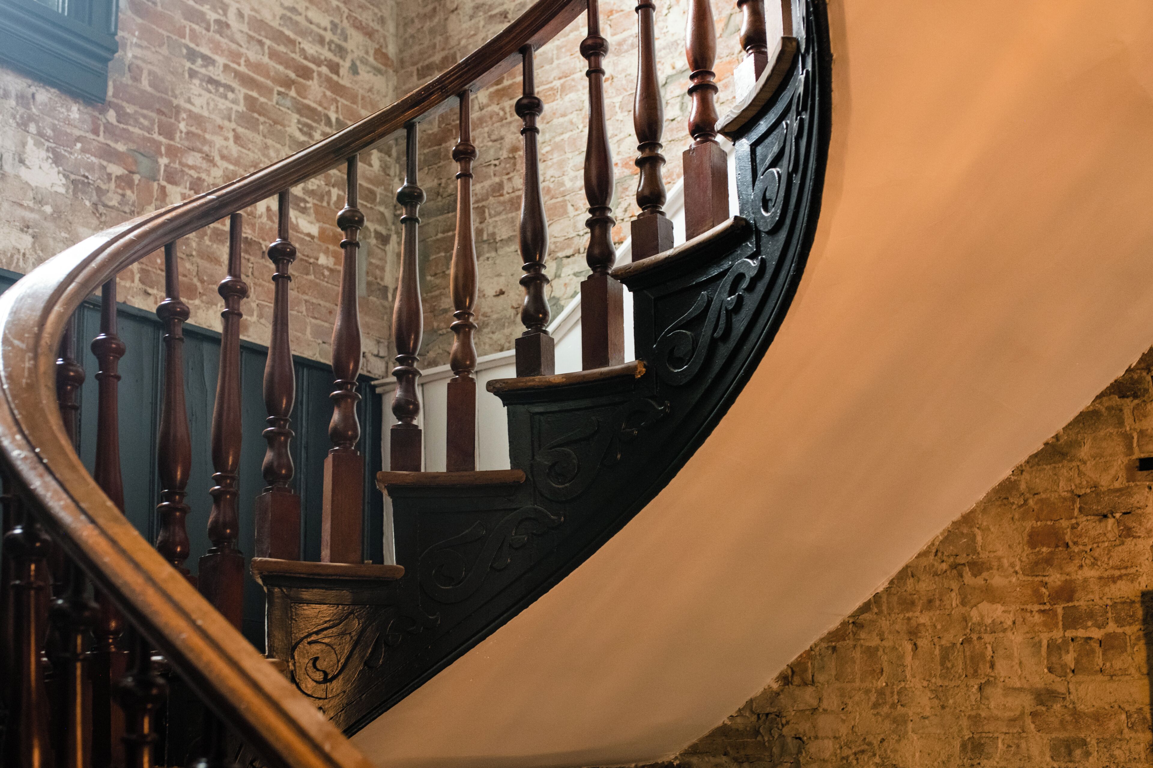 A spiral staircase with wooden balusters and a black handrail, leading to a brick wall.