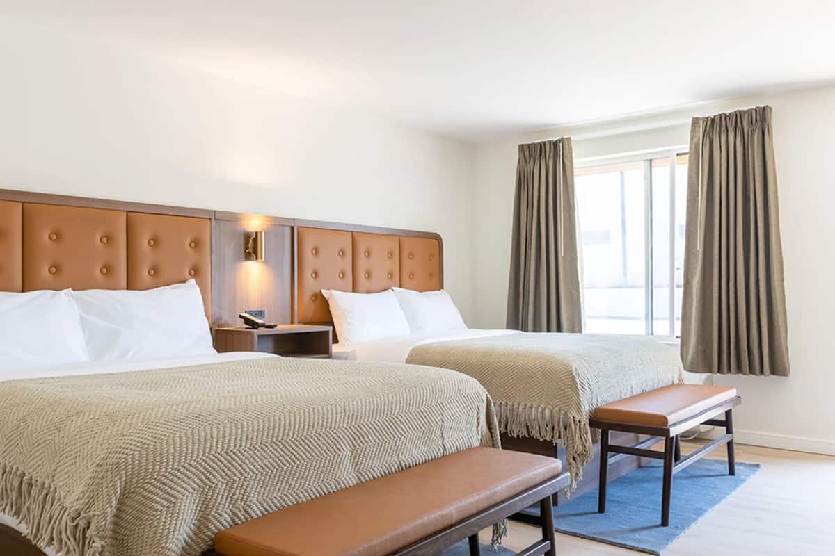 A hotel room with two beds, a bench, a nightstand, and a window with curtains.