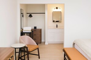 A hotel room with a bed, a small table with a chair, a vanity with a mirror, and a wardrobe.