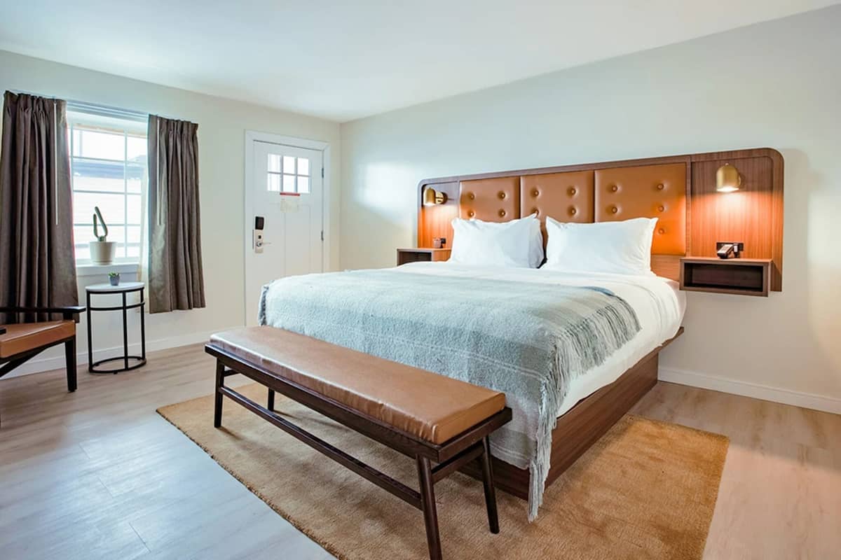 A hotel room with a bed, bedside tables, a bench, a small table, and a window with curtains.