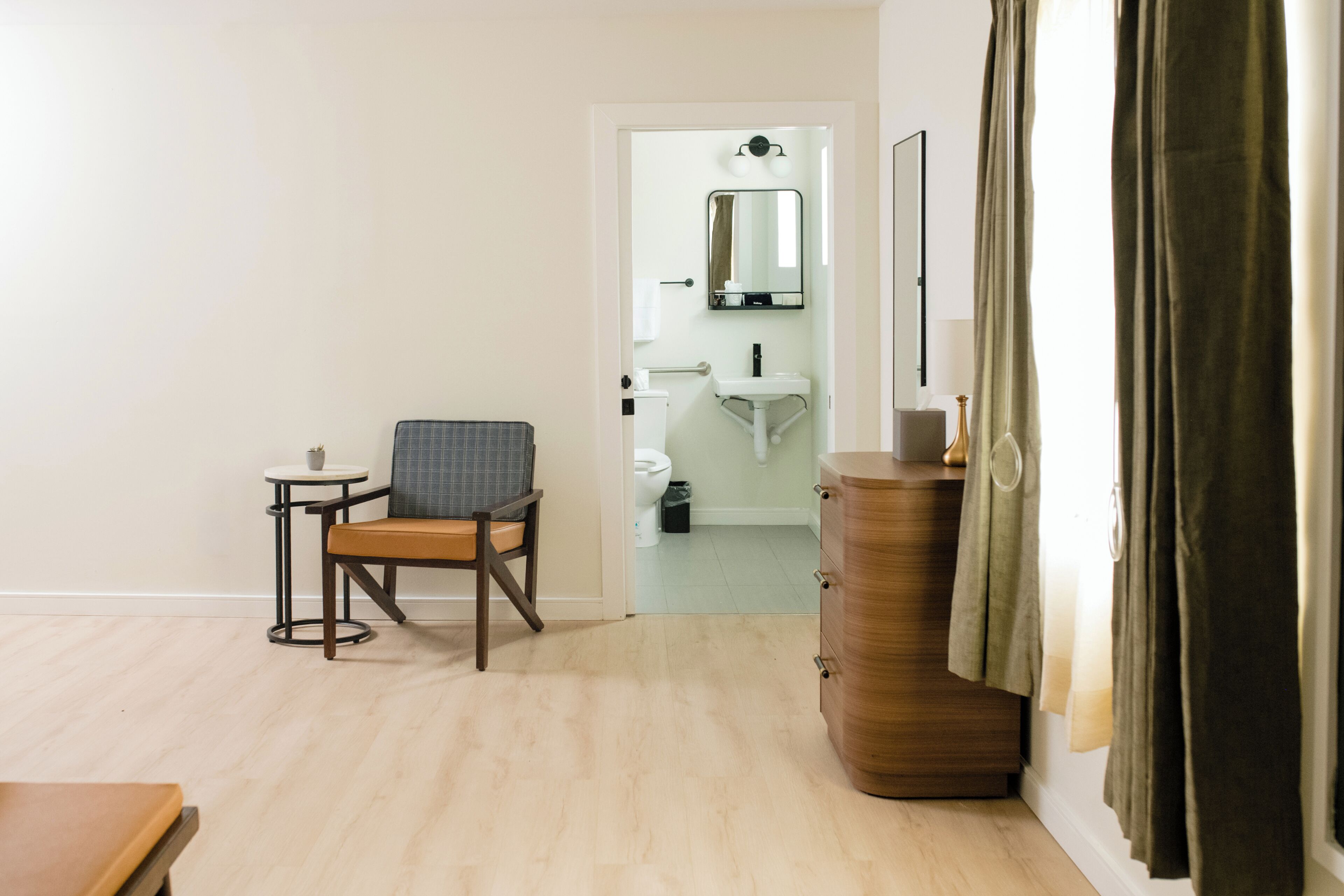 A hotel room with a wooden dresser, a chair, a small table, and a bathroom visible through an open door.