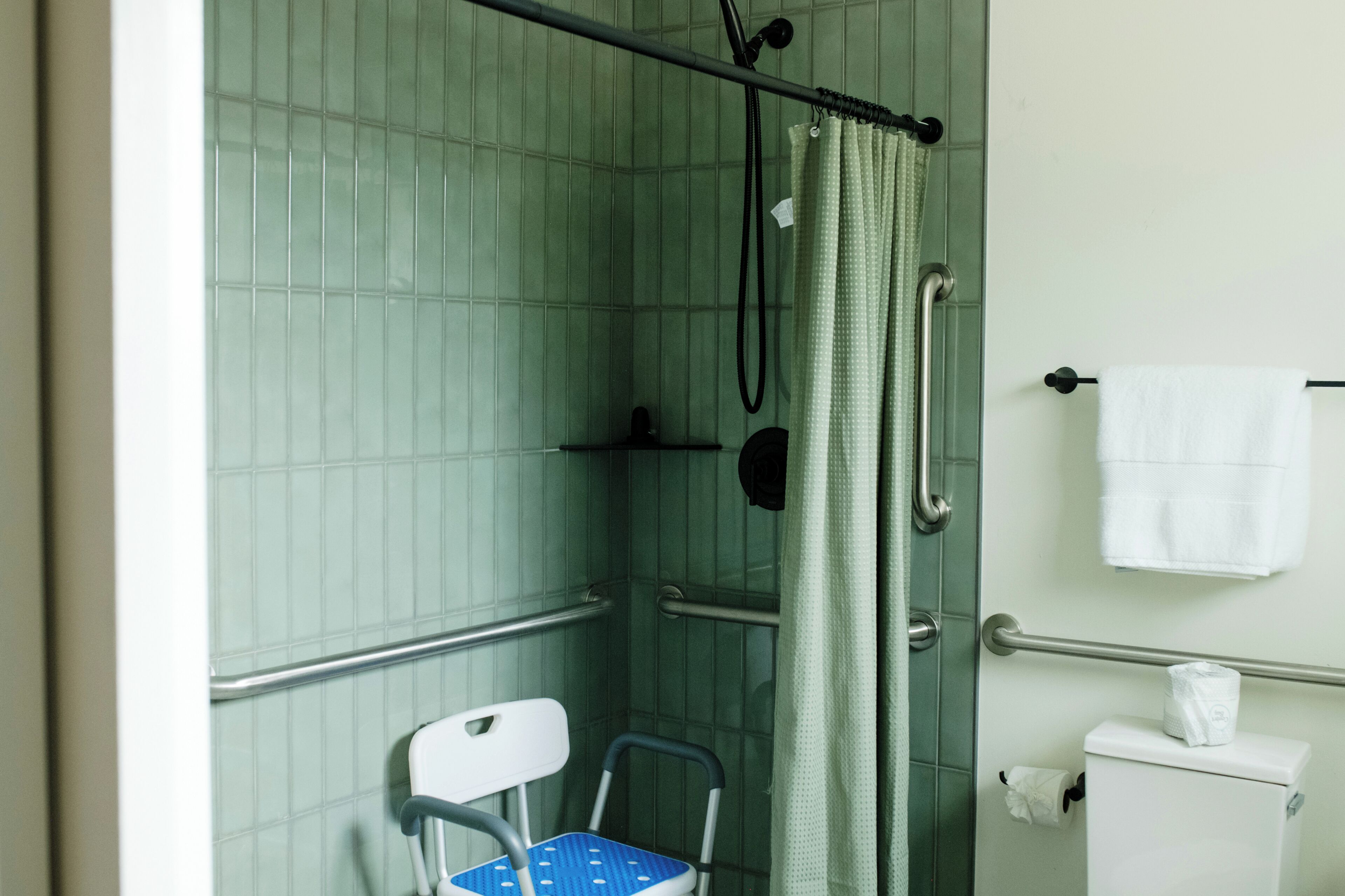 Baño con ducha, inodoro, barra de apoyo y silla de ruedas accesible.
