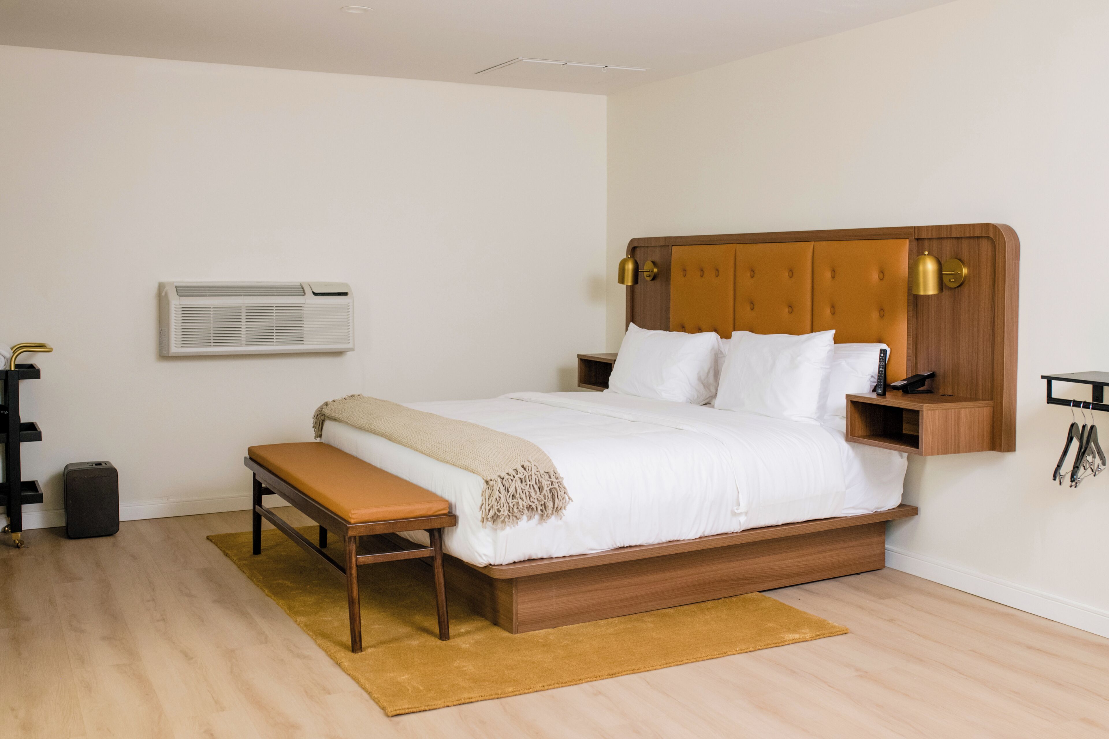 A bedroom with a wooden bed, a bench, a bedside table, an air conditioning unit, and a wall-mounted shelf.