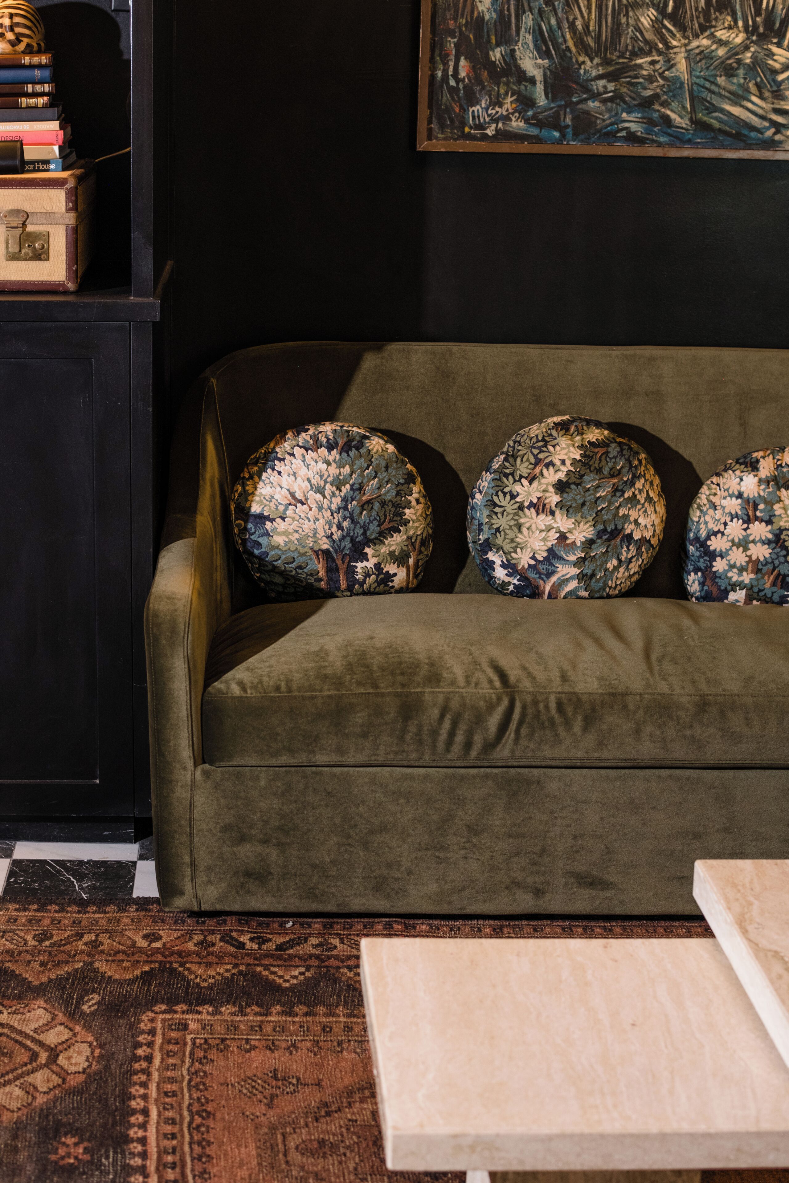 A green sofa with floral-patterned cushions, a dark-colored cabinet with books, and a painting on the wall.