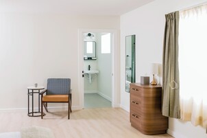 A room with a chair, a small table, a dresser, a mirror, and a bathroom visible through an open door.