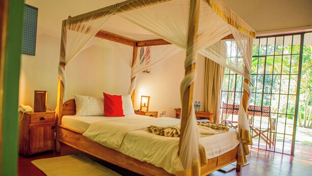 A four-poster bed with a canopy, a bedside table with a lamp, and a view of the outdoors through large windows. - Keeney House hotel  (Arusha)
