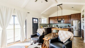 A spacious living area with a large window, a kitchen with wooden cabinets, a dining table, and a black leather sofa. - Charming 1-bedroom cottage in serene countryside (Blum)