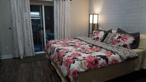 Un dormitorio con una cama, ropa de cama floral, una mesita de noche, una lámpara y vistas a un balcón.