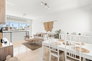 Une cuisine moderne avec des meubles blancs, un coin repas avec des tables et des chaises blanches, et une grande fenêtre donnant sur un paysage urbain.