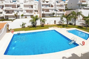 Une piscine dotée de deux échelles, entourée d’un bâtiment blanc agrémenté de balcons et de palmiers.