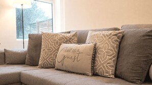 En beige hjørnesofa med mønstrede puder og en pude med teksten "BE OUR Guest".