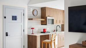 A compact kitchen with wooden cabinets, a microwave, a red kettle, and a wicker chair.