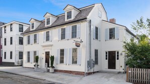 A white two-story building with a flat roof, multiple windows with shutters, and a small front porch with potted plants.