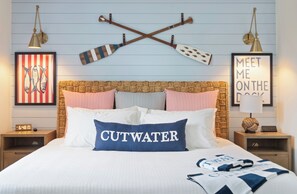 Habitación de hotel ordenada con una cama que incluye una almohada 'CUTWATER', almohadas a rayas y un cabecero decorativo. Hay dos mesitas de noche con lámparas y un cuadro enmarcado en la pared.