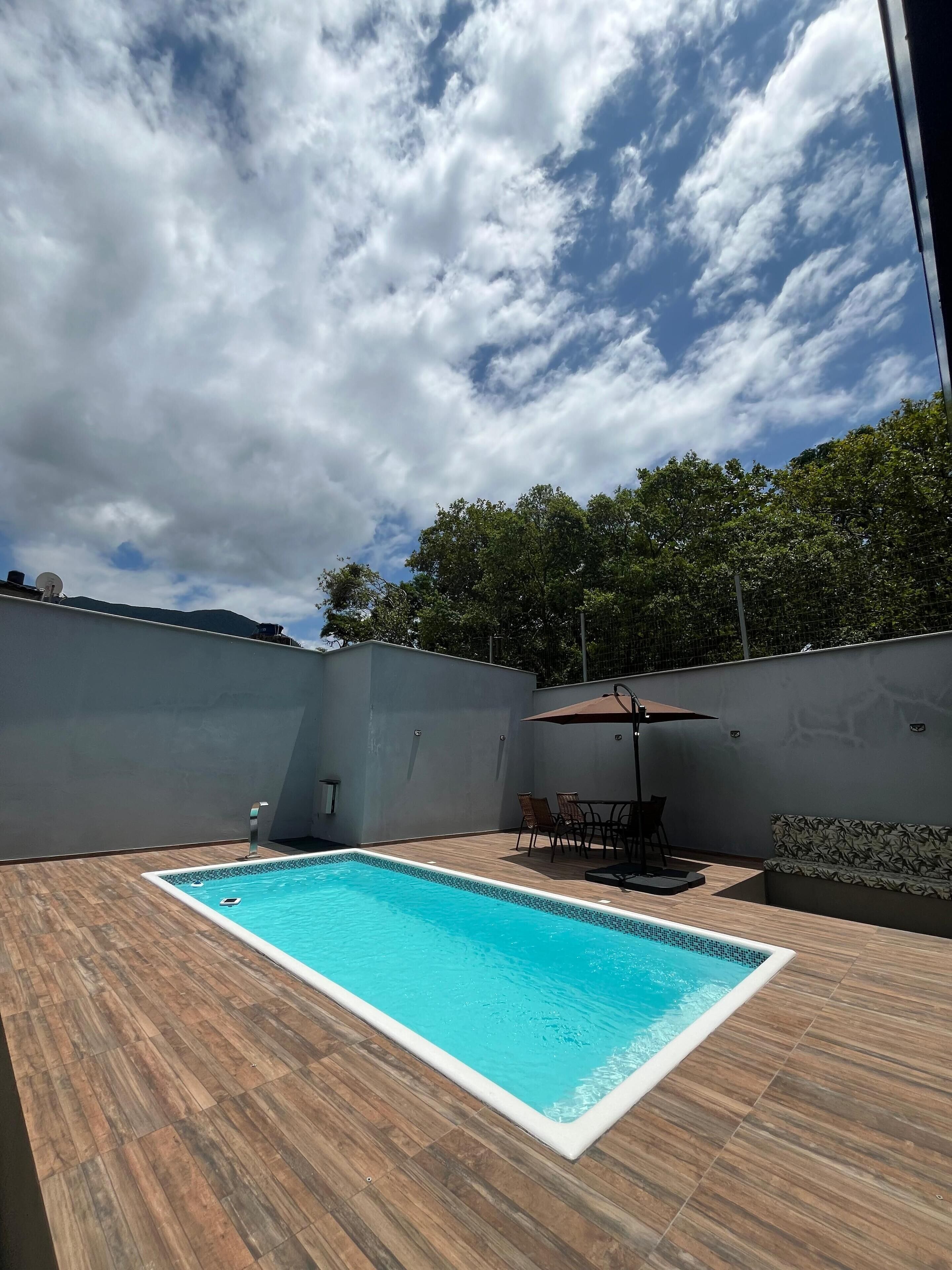 A rectangular swimming pool with a wooden deck, surrounded by a low wall and a covered seating area with chairs and a table.