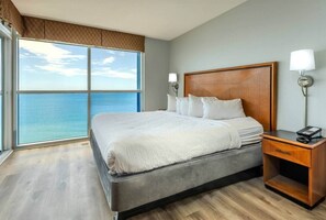 A hotel room with a large bed, a wooden headboard, two bedside lamps, a nightstand, and a view of the ocean. - Beachfront Boardwalk Condo (Myrtle Beach)