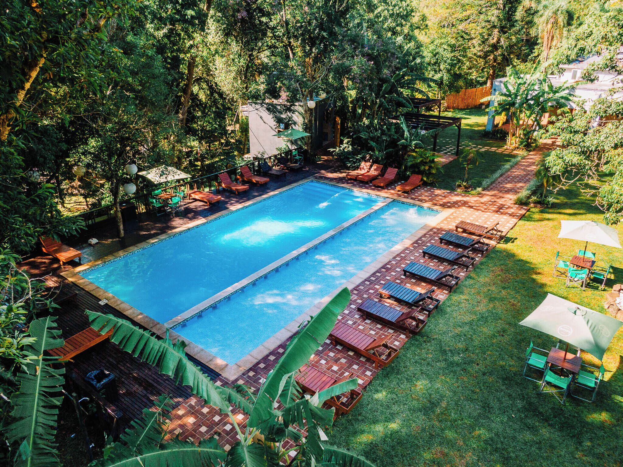 A rectangular swimming pool with a tiled deck, surrounded by lounge chairs and umbrellas, set in a lush garden with dense foliage.