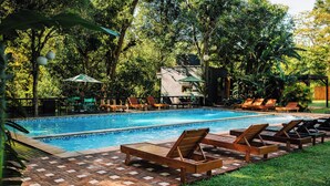 A rectangular swimming pool with a tiled edge, surrounded by a brick patio. There are wooden lounge chairs aligned along the pool's edge, and a small building with a green roof is visible in the background.