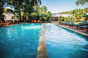 En swimmingpool med solstolar och parasoller, omgiven av frodig grönska och en byggnad i bakgrunden.